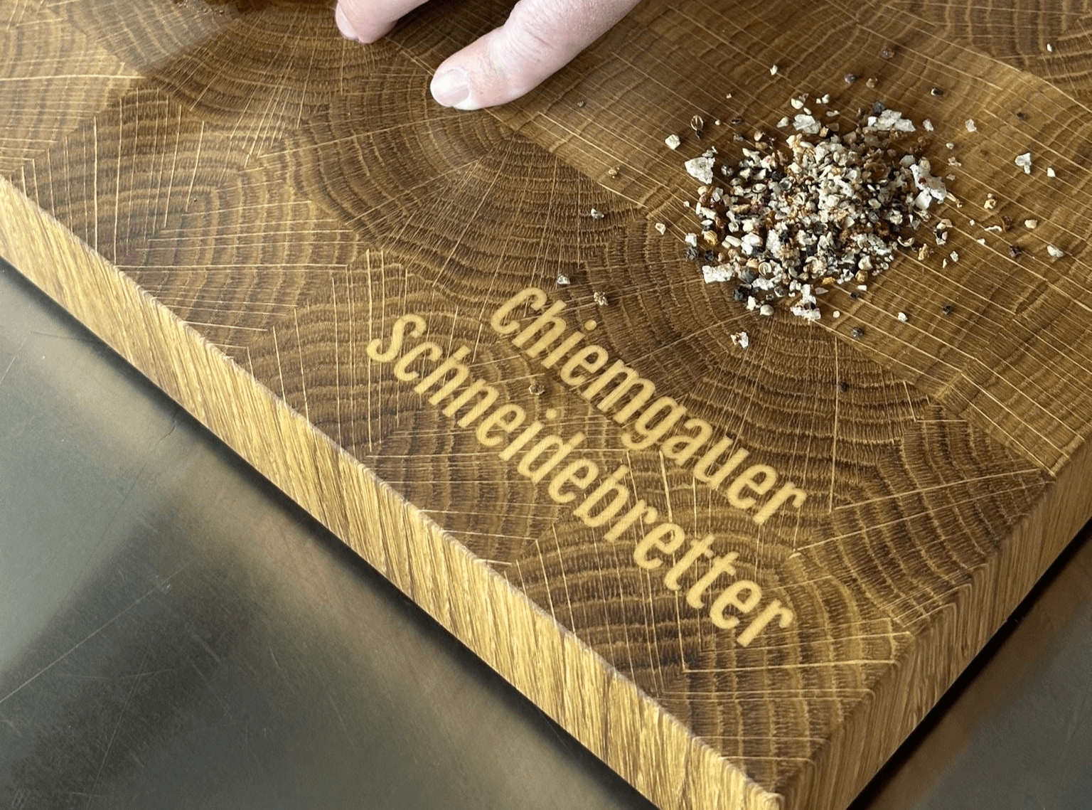 Chiemgauer Schneidebrett mit dem 1 cm tief eingravierten Schriftzug ‚Chiemgauer-Schneidebretter‘, aus Eiche und Ahorn gefertigt. Auf dem Brett wird ein Steak geschnitten, während daneben eine kleine Portion Pfefferkörner liegt